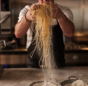 A chef lifting spaghetti out of a bowl of flour with flour visible in the air.