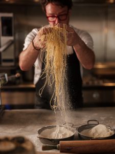 A chef lifting up hands full of spaghetti hanging down with flour releasing out.