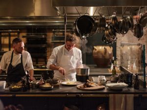 Two chefs at the pass one adding polenta to a plate with boards and pans around them and pans hanging from hooks above them.