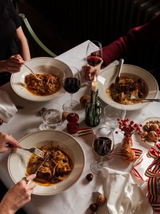Three people eating bowls of lasagne at a table with glasses of red wine.