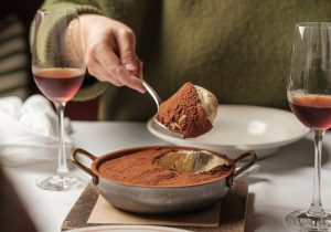A sharing bowl of tiramisu with a spoon full hovering over it and two glasses of dessert wine.