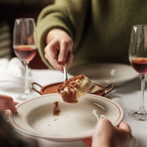 A person holding a spoon full of tiramisu over a plate with glasses of dessert wine.