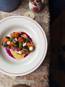 A colourful dish on a windowsill with a piece of bruschetta covered in vegetables purees and ricotta.