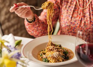 A bowl of tagliolini with Durslade Farm lamb ragu & cacioricotta with a spoon full of pasta and a glass of red wine.