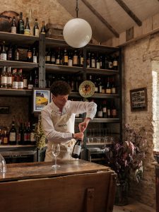 A person opening a bottle of red wine with a corkscrew in front of shelves full of wine.