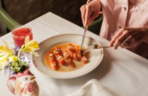 A plate of cured trout on a table with a white table cloth, a negroni and a jar of spring flowers.