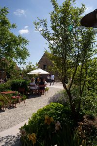 Outdoor garden restaurant with diners seated at tables under white umbrellas on gravel pathways. Lush landscaping includes mature trees, flowering plants, and lavender borders. Stone building visible in background under blue sky with white clouds.