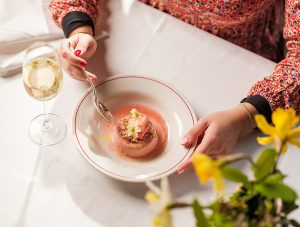 Rhubarb panna cotta on table with a glass of white wine and a spoon in a hand ready to dig in.