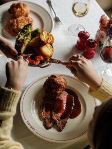 Woman sat at a table in Da Costa restaurant adding roasted carrots, greens, and roast potatoes to a plate of Durslade Farm Beef Sirloin and a Yorkshire pudding.