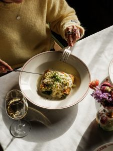 Woman about to tuck into baked Cannelloni with Westcombe ricotta, swiss chard, and cheddar sauce at Da Costa restaurant with a glass of white wine.