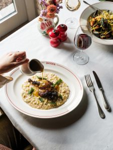Woman pouring sauce over a plate of leek risotto with confit and smoked duck leg.