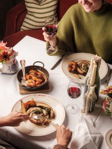 Guests eating Da Costa porchetta at the restaurant table surrounded by red wine, lit candles and a plate of roasted vegetables.