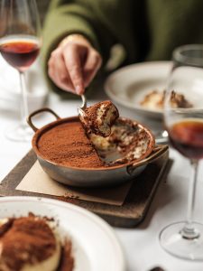Close-up of someone lifting a spoon of cocoa-dusted tiramisu from the dessert plate beside glasses of red wine on a restaurant table.