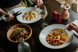 Two white dinner plates of seasonal dishes on a table, with two glasses of white wine