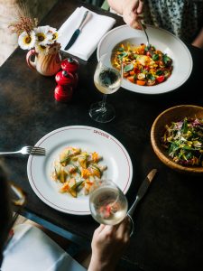 Two white dinner plates of seasonal dishes on a table, with two glasses of white wine
