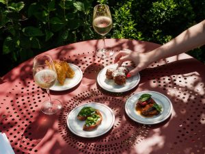 Four plates of different snacks, including pizzaiola arancini and pea and ricotta cicchetti, set on a red table outside with dappled sunlight beside two glasses of white wine.