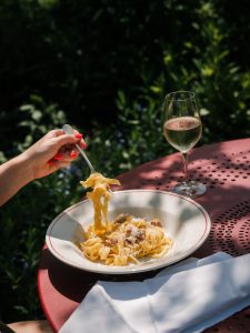 A woman tucking into a bowl of tagliatelle topped with guanciale and pecorino, set on a red outside table next to a white napkin and white glass of wine, with green bushes in the backdrop.